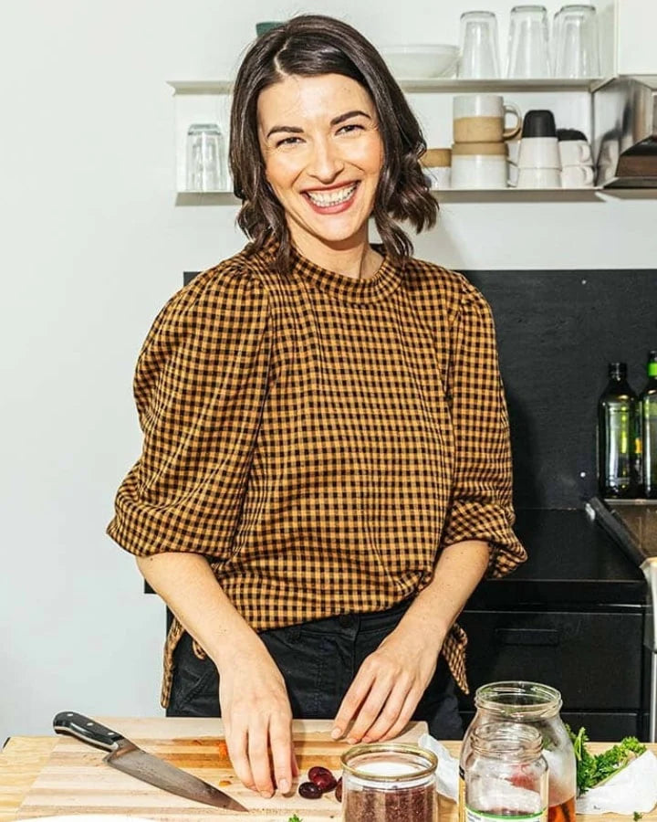 A woman in a brown checkered top stands smiling in a kitchen. She is preparing food on a wooden countertop with a jar and knife nearby. Shelves with cups and plates are visible in the background.